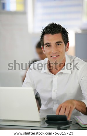 Portrait of a young man sitting at a desk typing on a calculator in front of a laptop computer