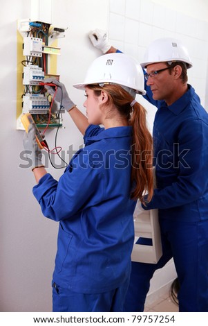 a young female electrician using an ammeter for checking an electricity meter and an older man watching her