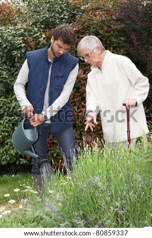 A gardener watering flowers in a garden and an elderly lady making comments as she watches him.