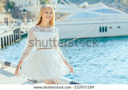 Smiling female in white dress on sea coast with yacht in the background