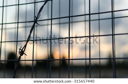Strong barbed wire fence and wire mesh barrier with blurred clouds as a background