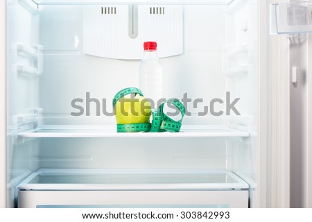 Green apple with measuring tape and a bottle of water on shelf of open empty refrigerator. Weight loss diet concept.
