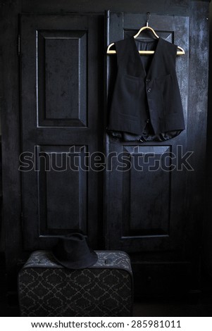 A waistcoat on a wooden hanger and a suitcase on a floor with a black hat on it