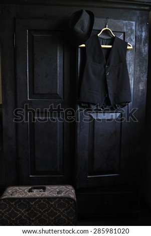 A waistcoat on a wooden hanger with  a hat on it and a suitcase on the floor
