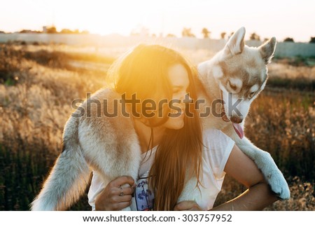 young caucasian female playing with her siberian husky puppy in the field during the sunset. Happy smiling girl having fun with puppy outdoors in beautiful light