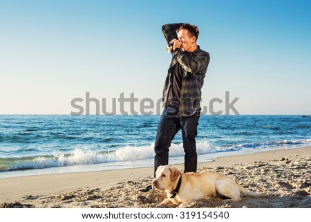 young caucasian male drinking coffee on beach while walking with dog during sunrise