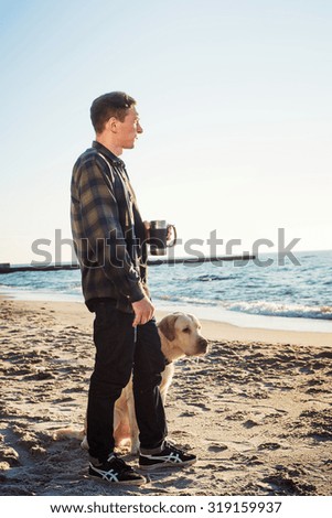 young caucasian male drinking coffee on beach while walking with dog during sunrise