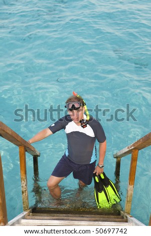 Man stands with equipment for Snorcel on steps of villa on piles on water.  Maldives.