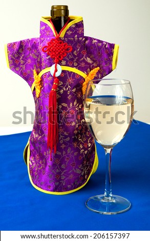 Bottle with Chinese white wine, decorated with a nice purple kimono with a pattern of flowers and yellow ribbon. A glass with wine next to it, all standing on a table with a blue table cloth