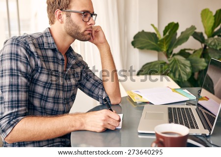 Young student looking at the screen of his new notebook while taking notes for his class that he is studying online, while drinking his morning cup of coffee