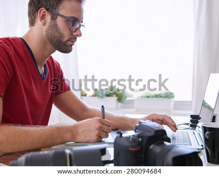 Handsome young man with a beard sitting at his glass desk in his home office busy editing images on his laptop with his camera visible in the foreground