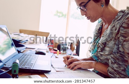 Mature business woman busy reading off of her printed list of employees while wearing glasses and sitting at her desk in her home office