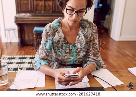 older business woman busy reading a text message on her cell phone while wearing glasses and sitting at her wooden desk
