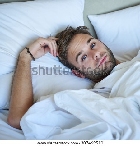 Confident young caucasian man lying in his white bedding scratching his head while looking at the camera