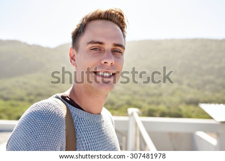Close up portrait image of a handsome young man busy smiling while looking straight at the camera and standing on the rooftop of his vacation home surrounded by nothing but bushes and trees