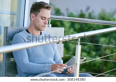 Image of a white young man sitting on his patio while looking up the stock markets on his tablet