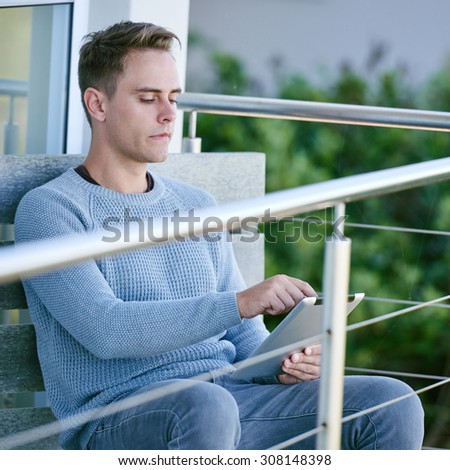 Image of a white young man sitting on his patio while looking up the stock markets on his tablet