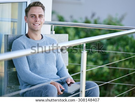 Young man smiling at camera while holding a tablet in his hands, and sitting on his patio outside his modern house
