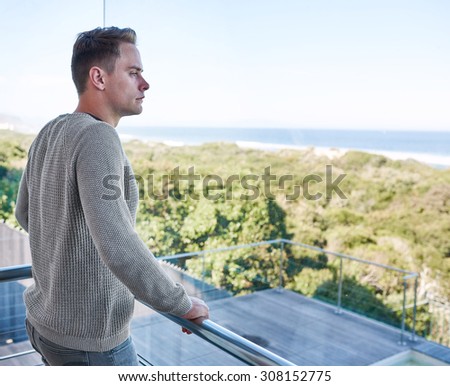 Young successful businessman standing on the balcony of his large luxurious beach house while looking out at the view of the ocean