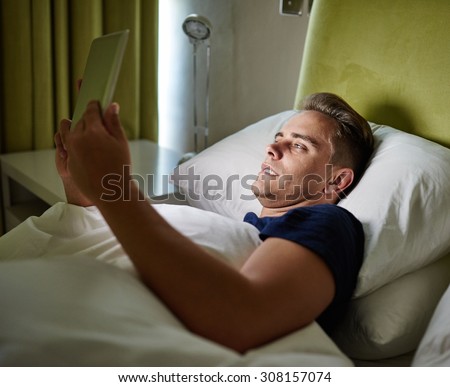 Young man busy lying in bed while using his tablet to search the internet for the weather report for the following day
