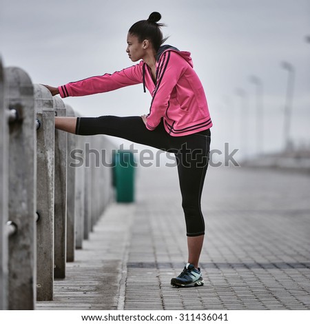 Square image of a young athlete busy stretching on the railing of the paved walkway along the ocean side