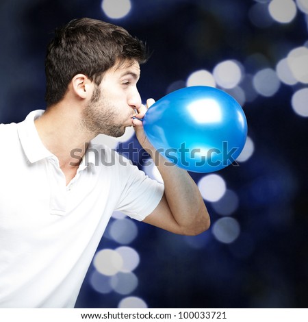 portrait of a young man inflating a blue balloon against a blue light background