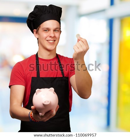 portrait of young cook man holding euro coin and piggy bank indoor