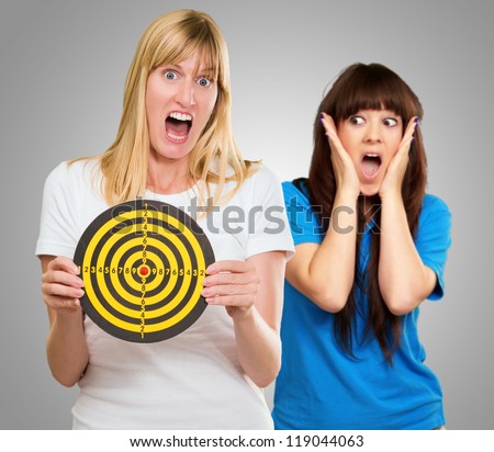 Woman Holding Dartboard In Front Of Shocked Woman On Gray Background