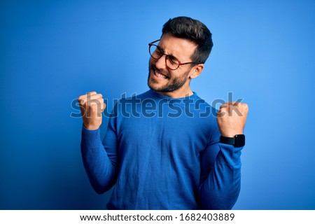 Photo of Young handsome man with beard wearing casual sweater and glasses over blue background very happy and excited doing winner gesture with arms raised, smiling and screaming for success. Celebration