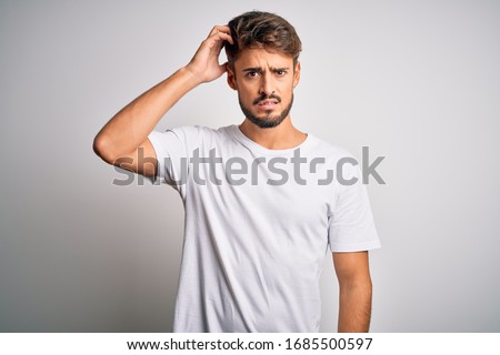 Young handsome man with beard wearing casual t-shirt standing over white background confuse and wonder about question. Uncertain with doubt, thinking with hand on head. Pensive concept.
