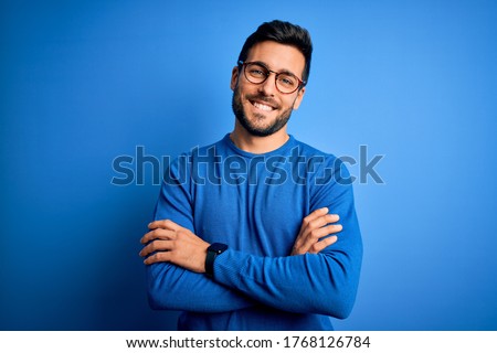 Photo of Young handsome man with beard wearing casual sweater and glasses over blue background happy face smiling with crossed arms looking at the camera. Positive person.