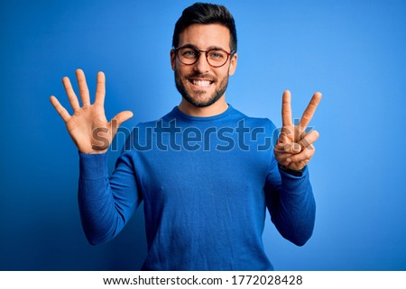 Young handsome man with beard wearing casual sweater and glasses over blue background showing and pointing up with fingers number seven while smiling confident and happy.