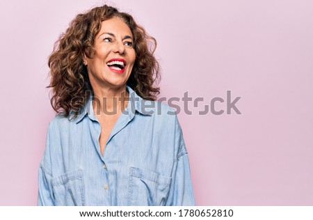 Middle age beautiful woman wearing casual denim shirt standing over pink background looking away to side with smile on face, natural expression. Laughing confident.