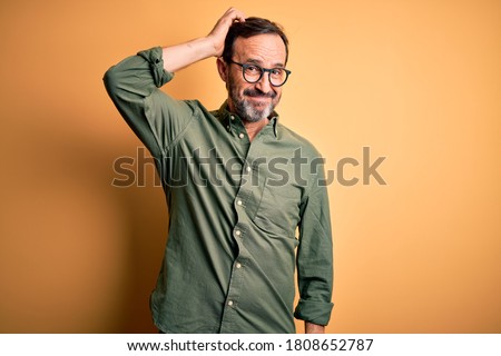 Middle age hoary man wearing casual green shirt and glasses over isolated yellow background confuse and wonder about question. Uncertain with doubt, thinking with hand on head. Pensive concept.
