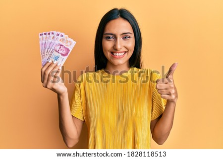 Young brunette woman holding 50 mexican pesos banknotes smiling happy and positive, thumb up doing excellent and approval sign  Foto stock © 
