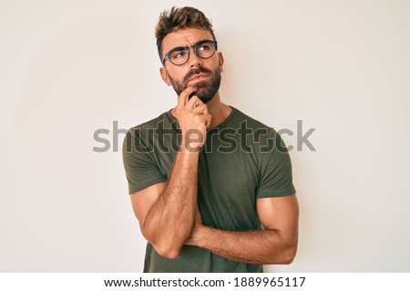 Young hispanic man wearing casual clothes and glasses serious face thinking about question with hand on chin, thoughtful about confusing idea 