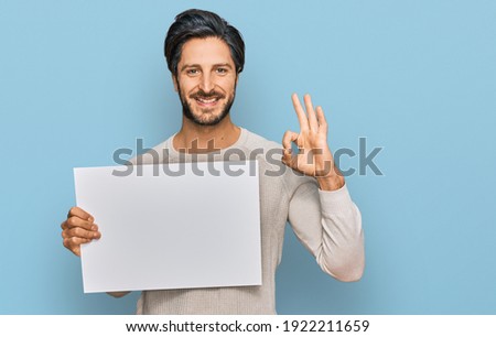 Young hispanic man holding blank empty banner doing ok sign with fingers, smiling friendly gesturing excellent symbol  Foto stock © 