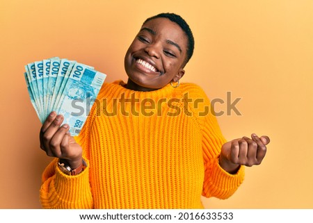 Young african american woman holding 100 brazilian real banknotes screaming proud, celebrating victory and success very excited with raised arm 