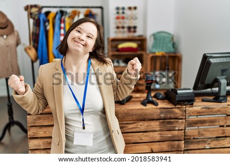 Young down syndrome woman working as manager at retail boutique very happy and excited doing winner gesture with arms raised, smiling and screaming for success. celebration concept. 