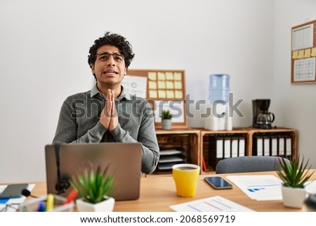 Young hispanic man wearing business style sitting on desk at office begging and praying with hands together with hope expression on face very emotional and worried. begging.  Foto d'archivio © 