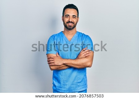 Handsome hispanic man with beard wearing blue male nurse uniform happy face smiling with crossed arms looking at the camera. positive person.  商業照片 © 