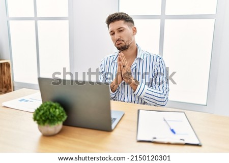 Young handsome man with beard working at the office using computer laptop begging and praying with hands together with hope expression on face very emotional and worried. begging.  Stockfoto © 