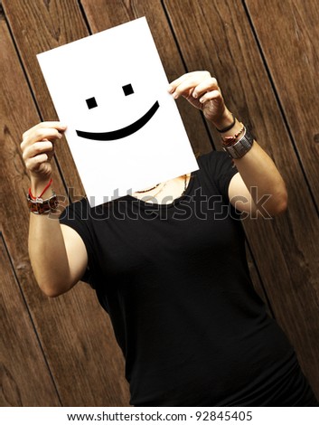 Woman showing a blank paper with a smile emoticon in front of her face against a wooden wall