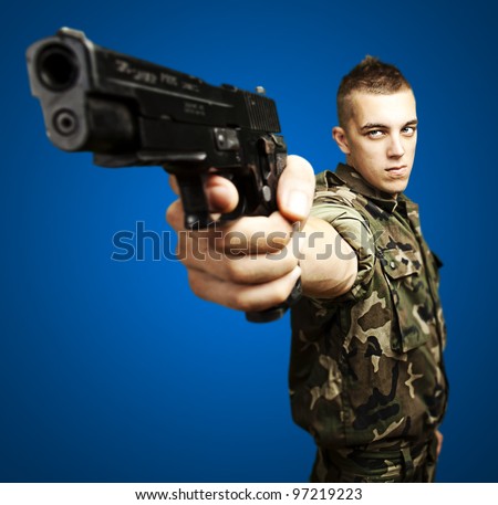 portrait of a caucasian soldier with a jungle camouflage pointing with a pistol over a blue background