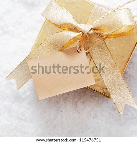 Gold Christmas gift box with blank paper label and decorative ribbon and bow standing on a bed of fresh winter snow