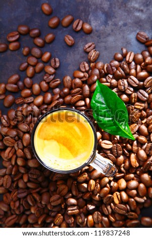 Overhead view of a cup of rich frothy coffee standing in scattered whole roasted coffee beans with a green leaf for decoration