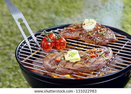 Tender steak topped with a curl of butter and herbs grilling outdoors on a portable barbecue with a pronged fork and tomatoes