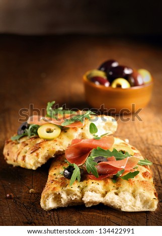 Serving of fresh traditional oven-baked Italian focaccia bread with ham, olives and rocket on a wooden table top with copyspace