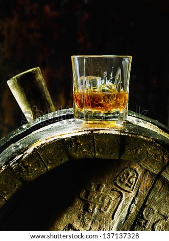 A glass of whiskey with ice on an old oak barrel using dramatic lighting.