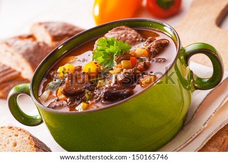 Close up view of a green metal pot filled with a tasty rich meaty stew with vegetables served with sliced baguette